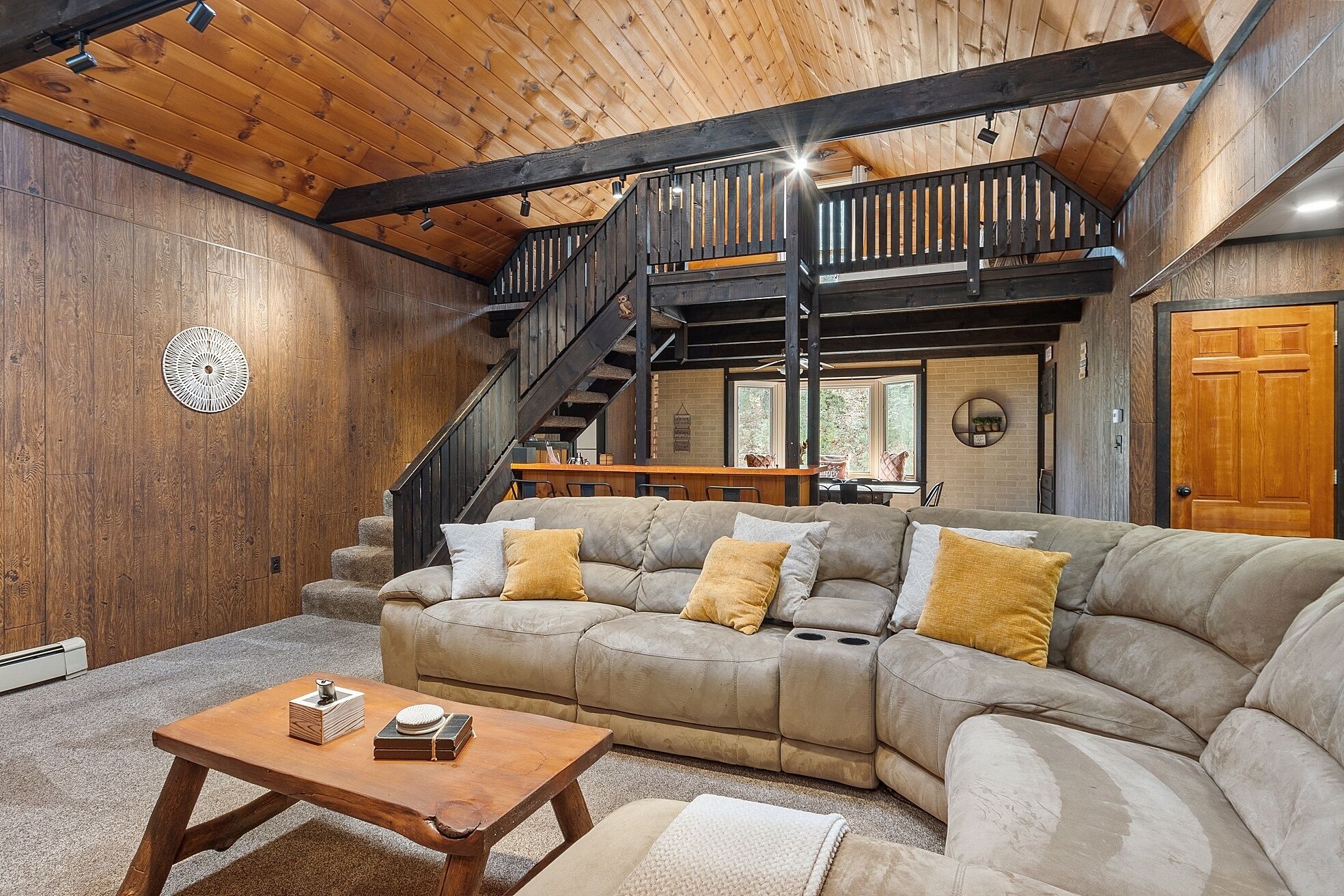 A cozy living room with a wooden ceiling, a large beige sectional sofa, a wooden coffee table, and a staircase leading to an upper level.