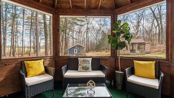 A cozy wooden cabin with wicker furniture, a glass coffee table, and a view of a small cabin in the distance.