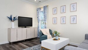 A modern living room with a flat-screen TV on a light-colored cabinet, a blue and white patterned rug, a grey sofa, and a coffee table with a vase of flowers.