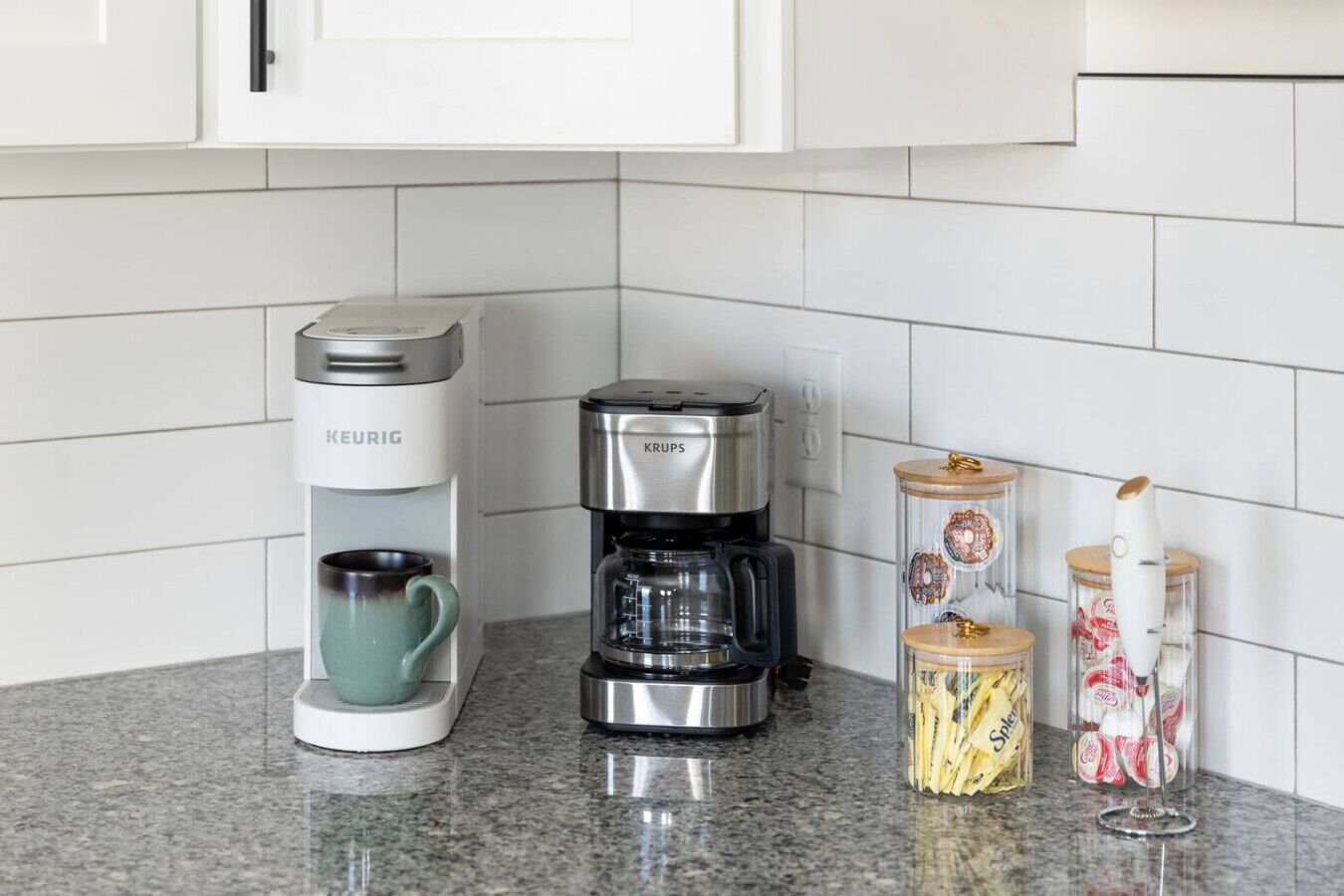 Une cuisine équipée d’une machine à café Keurig, d’une autre machine à café Krups et de deux bocaux en verre avec couvercles.