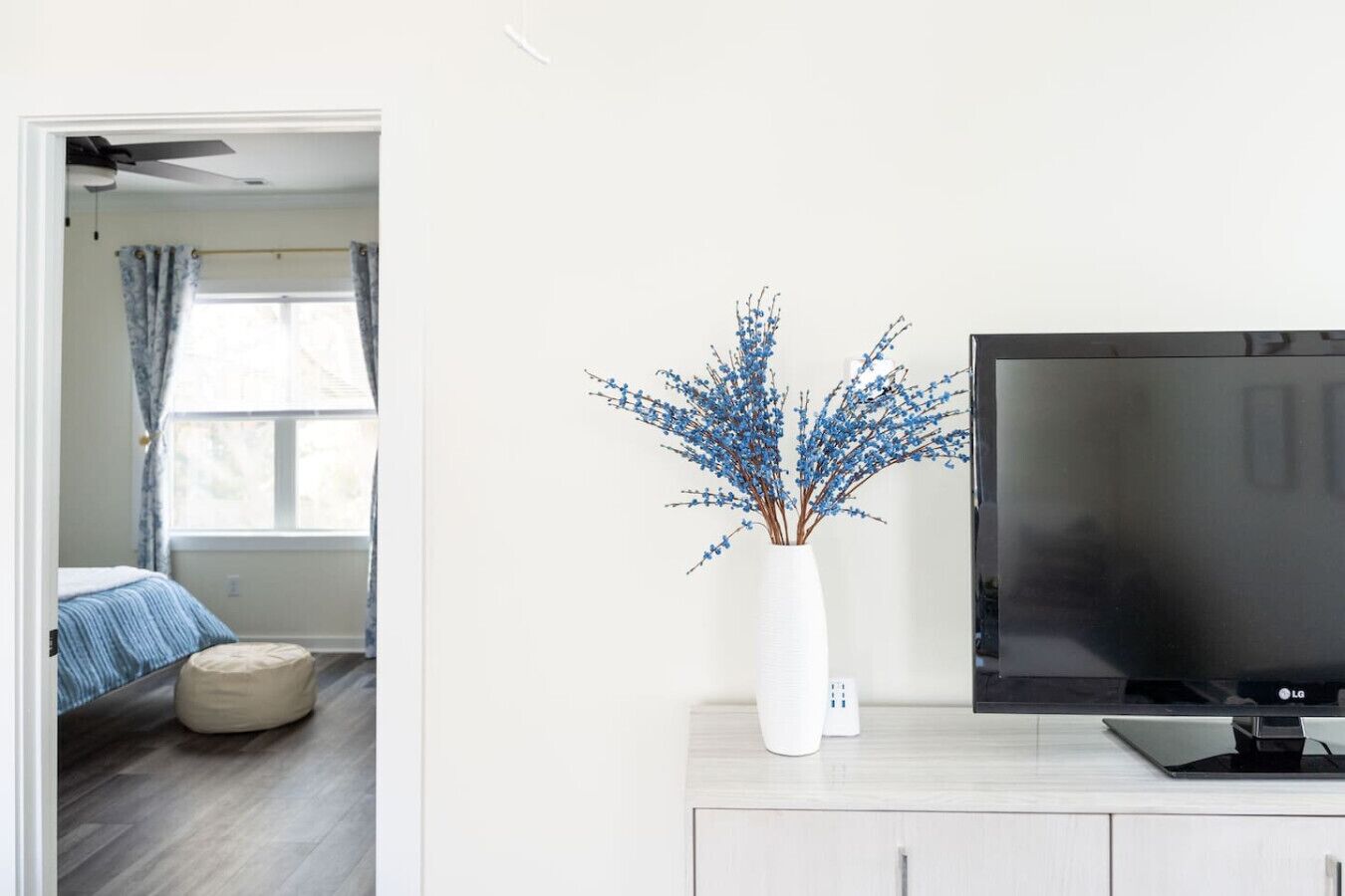 A modern living room with a flat-screen TV on a white cabinet, a vase with blue flowers, and a view into a bedroom with a bed and curtains.