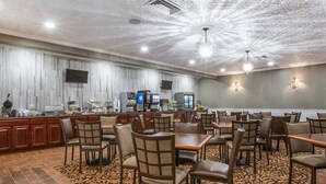 A hotel dining area with multiple tables and chairs, a buffet counter, and a television screen. - St. Lawrence Hotel  (Massena)