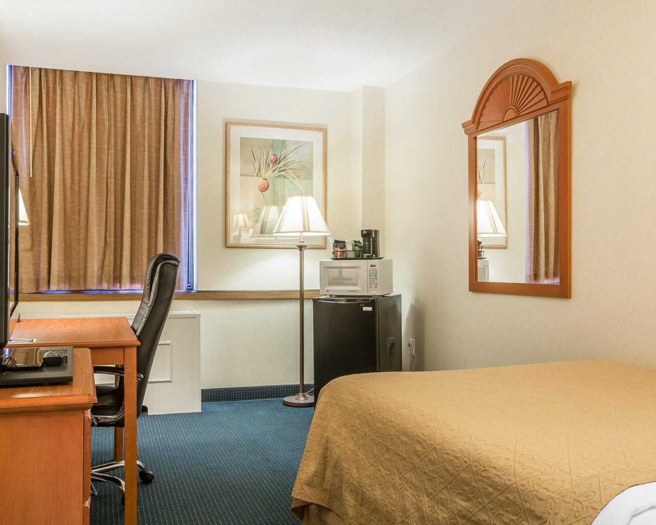 A hotel room with a bed, desk, chair, lamp, microwave, refrigerator, and a large mirror.