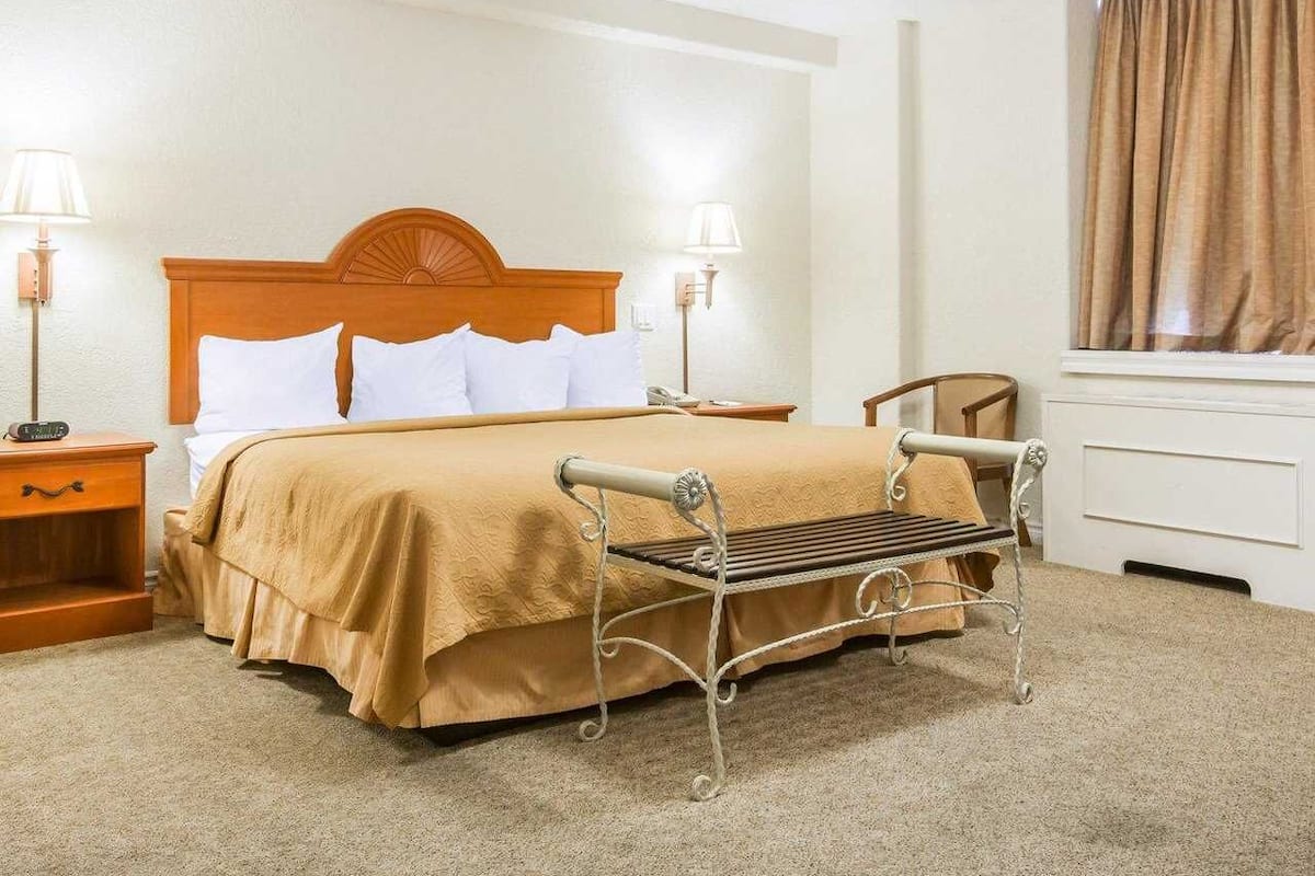 A hotel room with a bed, bedside tables, a nightstand, a chair, a dresser, and a window with curtains.