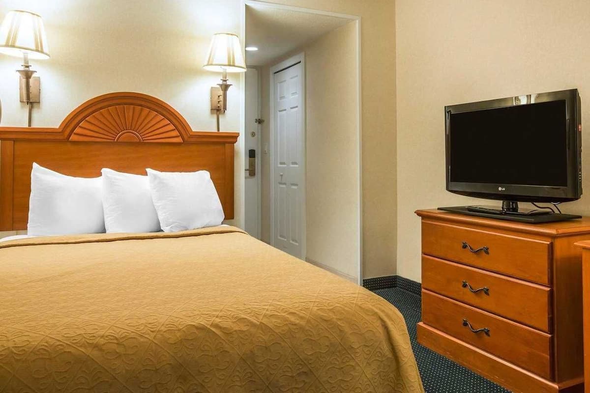 A hotel room with a bed, two wall-mounted lamps, a television on a dresser, and a door leading to another room.