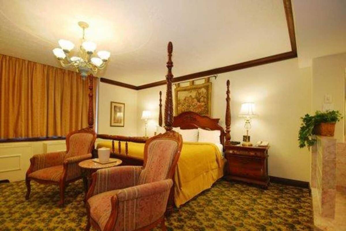 A hotel room with a four-poster bed, two armchairs, a small round table, and a nightstand with a lamp.