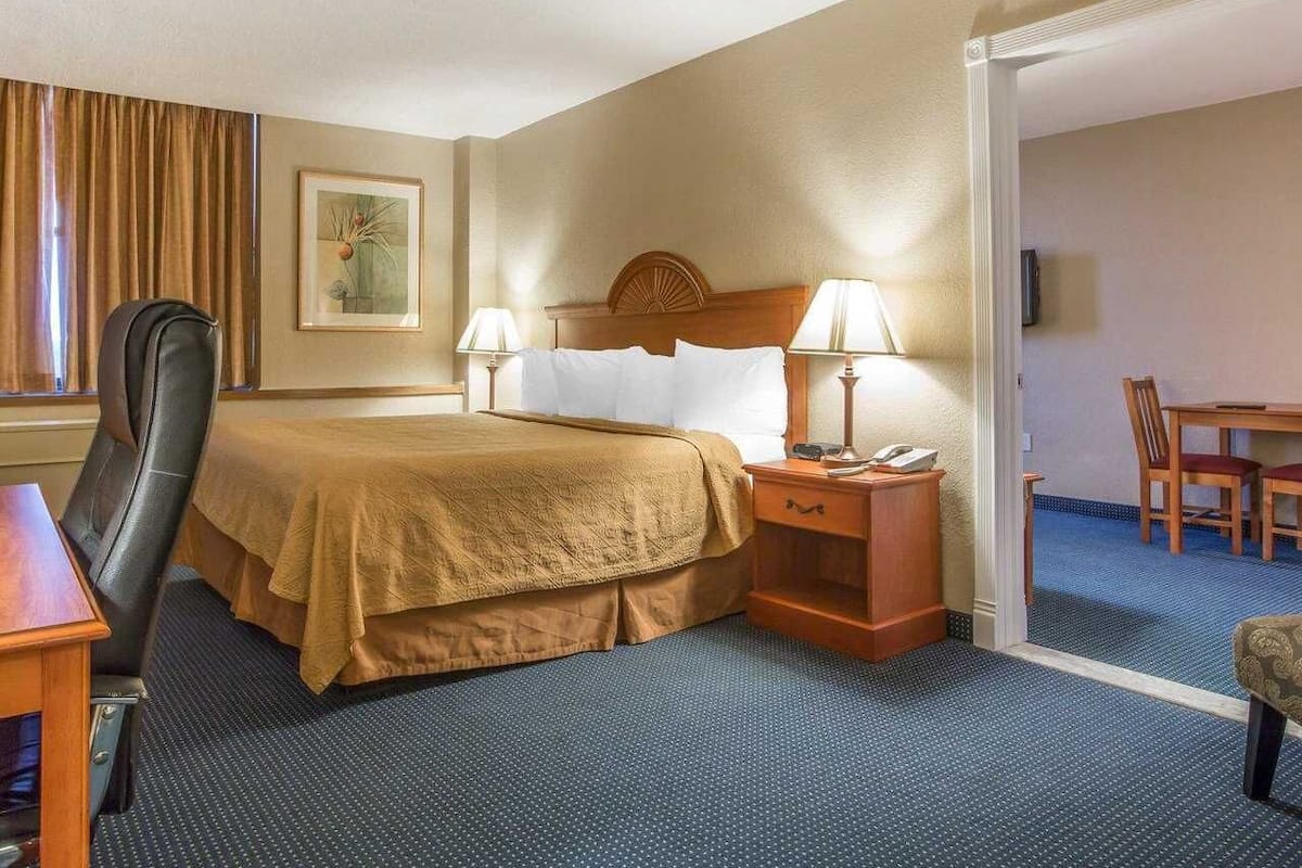 A hotel room with a bed, two bedside tables with lamps, a desk with a chair, a dining table with chairs, and a painting on the wall.