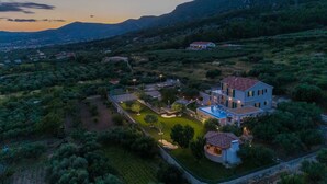 A large, two-story house with a swimming pool, surrounded by lush greenery and hills in the background.
