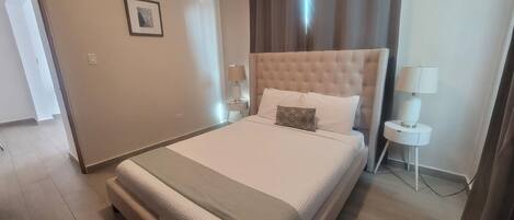 Une chambre d’hôtel avec un grand lit, une tête de lit, deux tables de chevet avec des lampes, un tableau encadré au mur et une porte donnant sur une autre pièce.