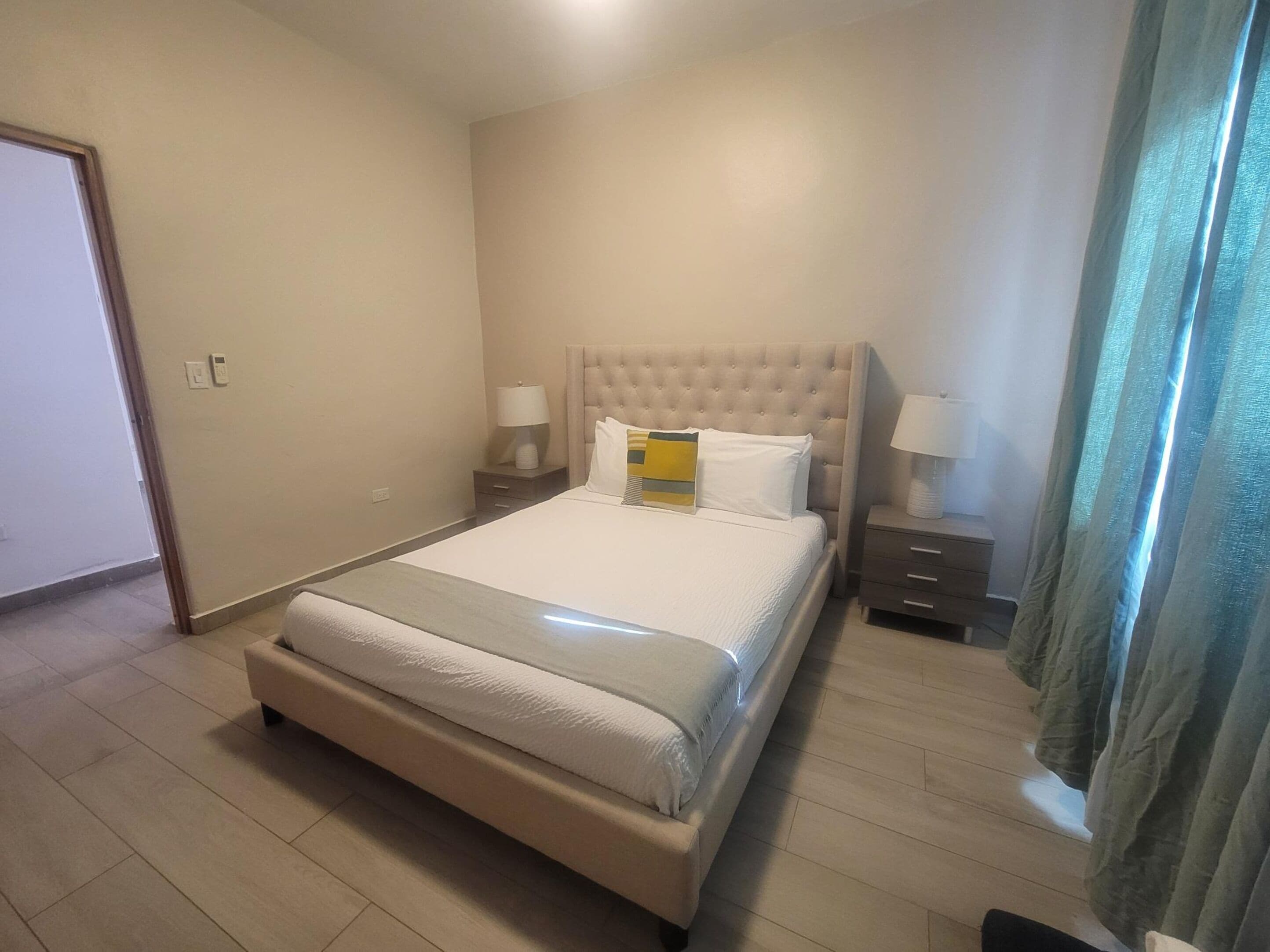 Une chambre d’hôtel avec un grand lit, deux tables de chevet avec des lampes, un plancher en bois et une porte située à gauche.
