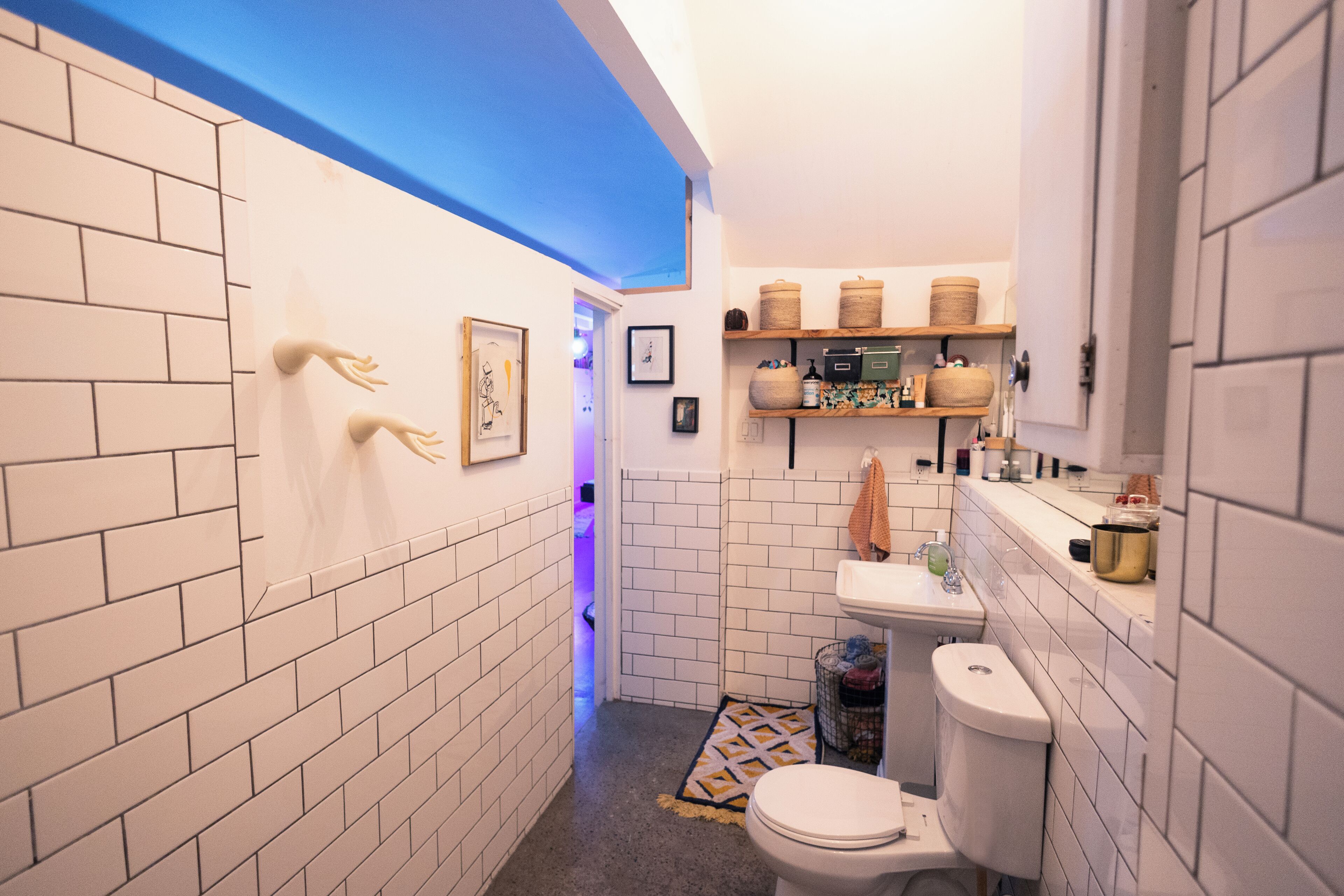 A narrow bathroom with white tiled walls, a blue ceiling, and a white sink with shelves above.