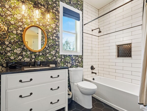 A bathroom with a black floral wallpaper, a white sink with black handles, a white toilet, and a white bathtub with a hexagonal window.