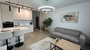 A modern kitchen with white cabinets, a dining area with a white table and chairs, a grey sofa, and a wooden coffee table.