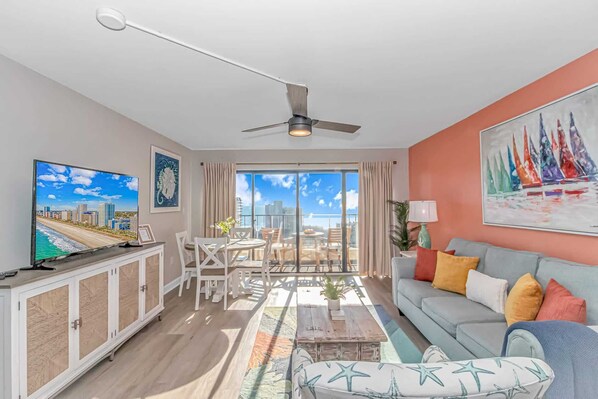 A living room with a ceiling fan, a television, a dining area with chairs and a table, a sofa with colorful cushions, and a large window with a view of the city.