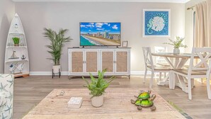 Een woonkamer met een televisie, een eettafel, stoelen, een kast en sierplanten.