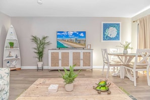 A living room with a TV, a dining table, chairs, a cabinet, and decorative plants. - Ocean Villas 902 (Myrtle Beach)