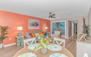 A living room with a ceiling fan, a dining table with a floral centerpiece, and a colorful mural on the wall.