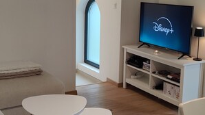 Ein modernes Wohnzimmer mit einem Flachbildfernseher, der Disney+ anzeigt, einem weißen Sofa, zwei kleinen Tischen und einem Holzboden.