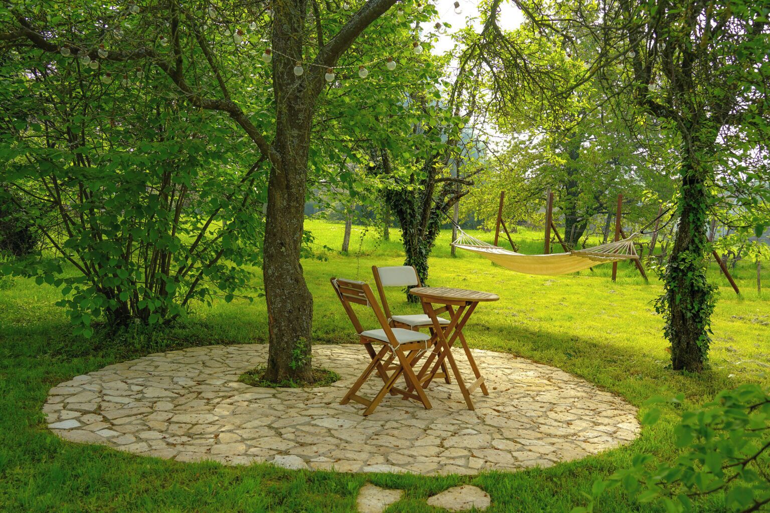A circular stone patio with a wooden table and chairs, surrounded by lush greenery and trees.