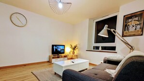 En moderne stue med et fladskærms-tv, et hvidt sofabord, en grå sofa, trægulv, et hvidt vægur og et vindue med et plakat.