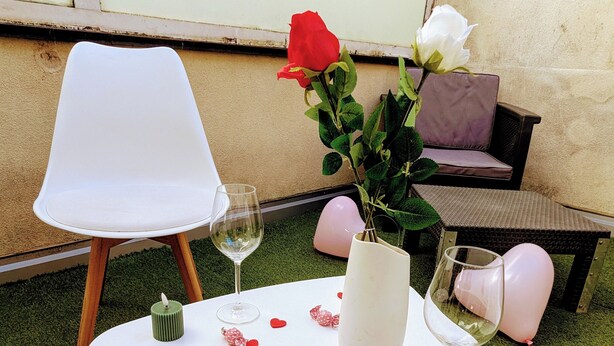 Et lille udendørs siddeområde med et hvidt bord, to stole, en vase med blomster og spredte hjerteformede dekorationer.