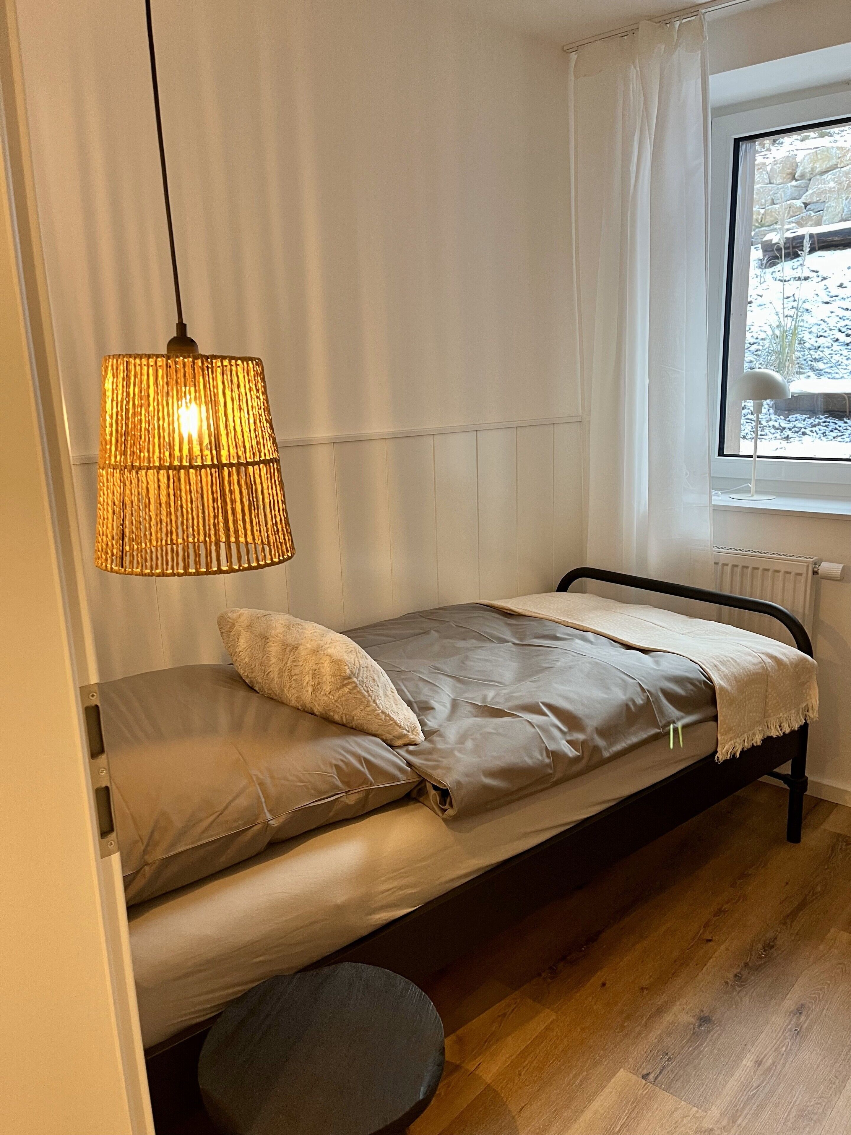 A small, single-bed room with a sloped ceiling, a window with curtains, and a hanging lamp.