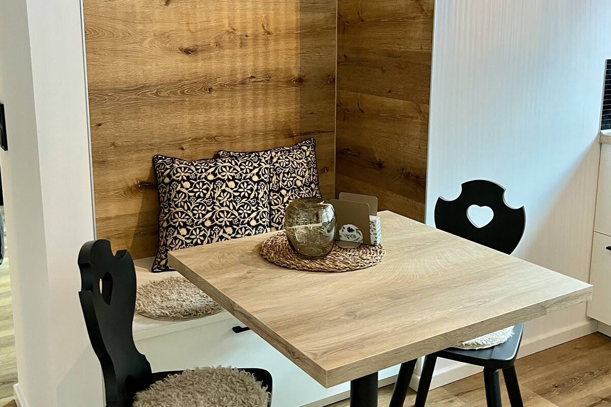 Un comedor con una mesa de madera, dos sillas, un jarrón decorativo y cojines estampados.