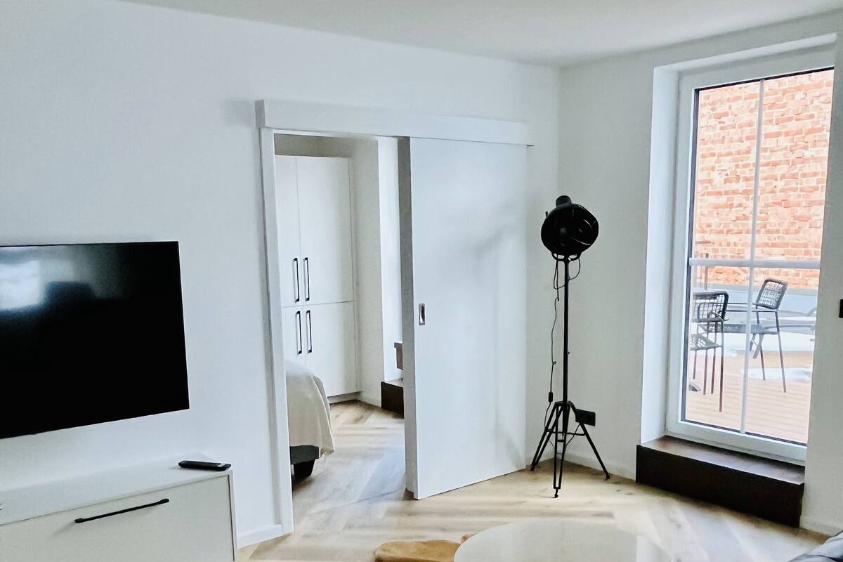 Una sala de estar moderna con televisor de pantalla plana, un sofá de cuero negro, una mesa de centro blanca, un tapete mullido y un ventanal con vistas a un edificio de ladrillo.
