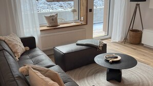 A modern living room with a dark leather sofa, a round coffee table, and a view of a snowy outdoor area through a large window. - Wyzenborne (Sontra)