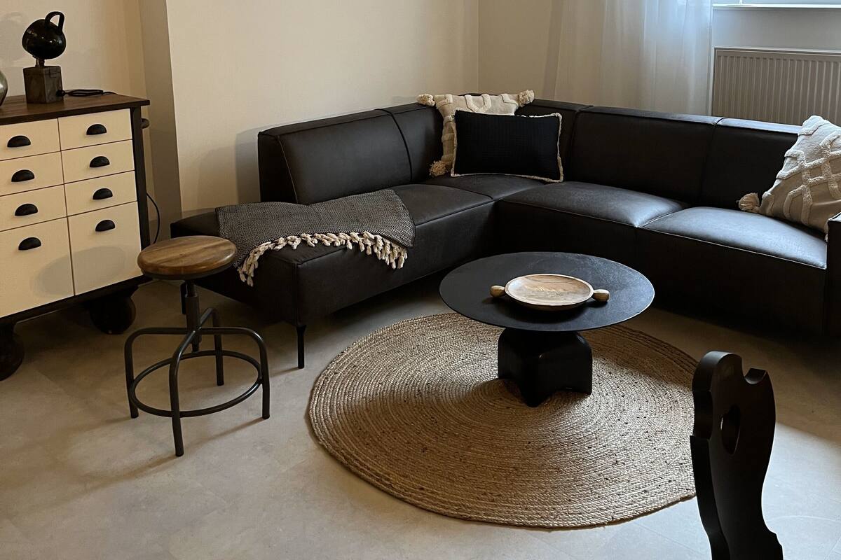 Una sala de estar moderna con un sofá de cuero oscuro, una mesa redonda de centro, un taburete pequeño y un mueble blanco con cajones.