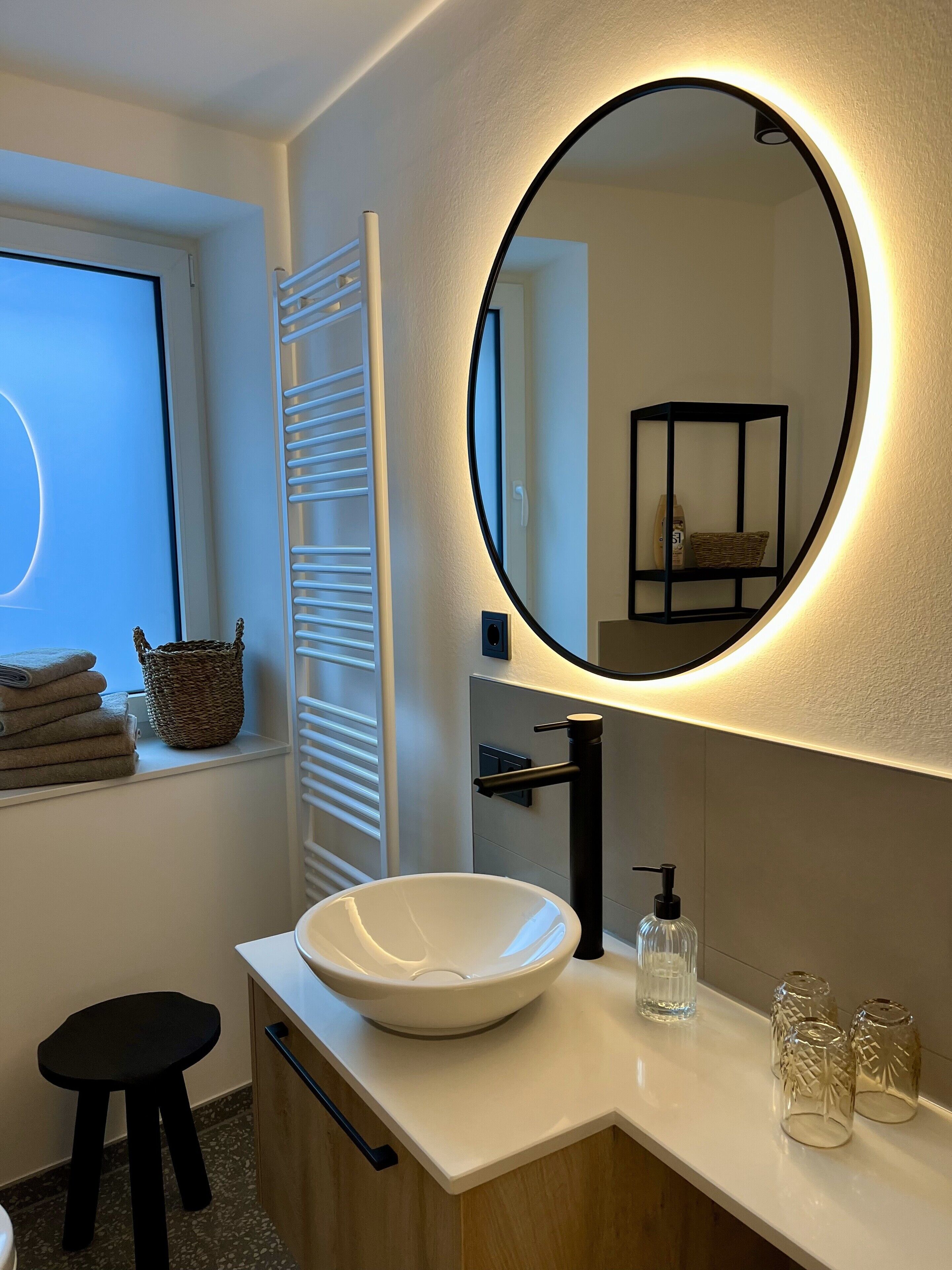 Un baño moderno con un espejo redondo, lavamanos blanco y una ventana con persianas.