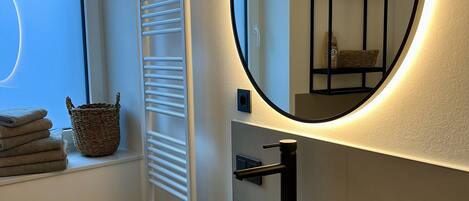 Un baño moderno con un espejo redondo, lavamanos blanco y una ventana con persianas.