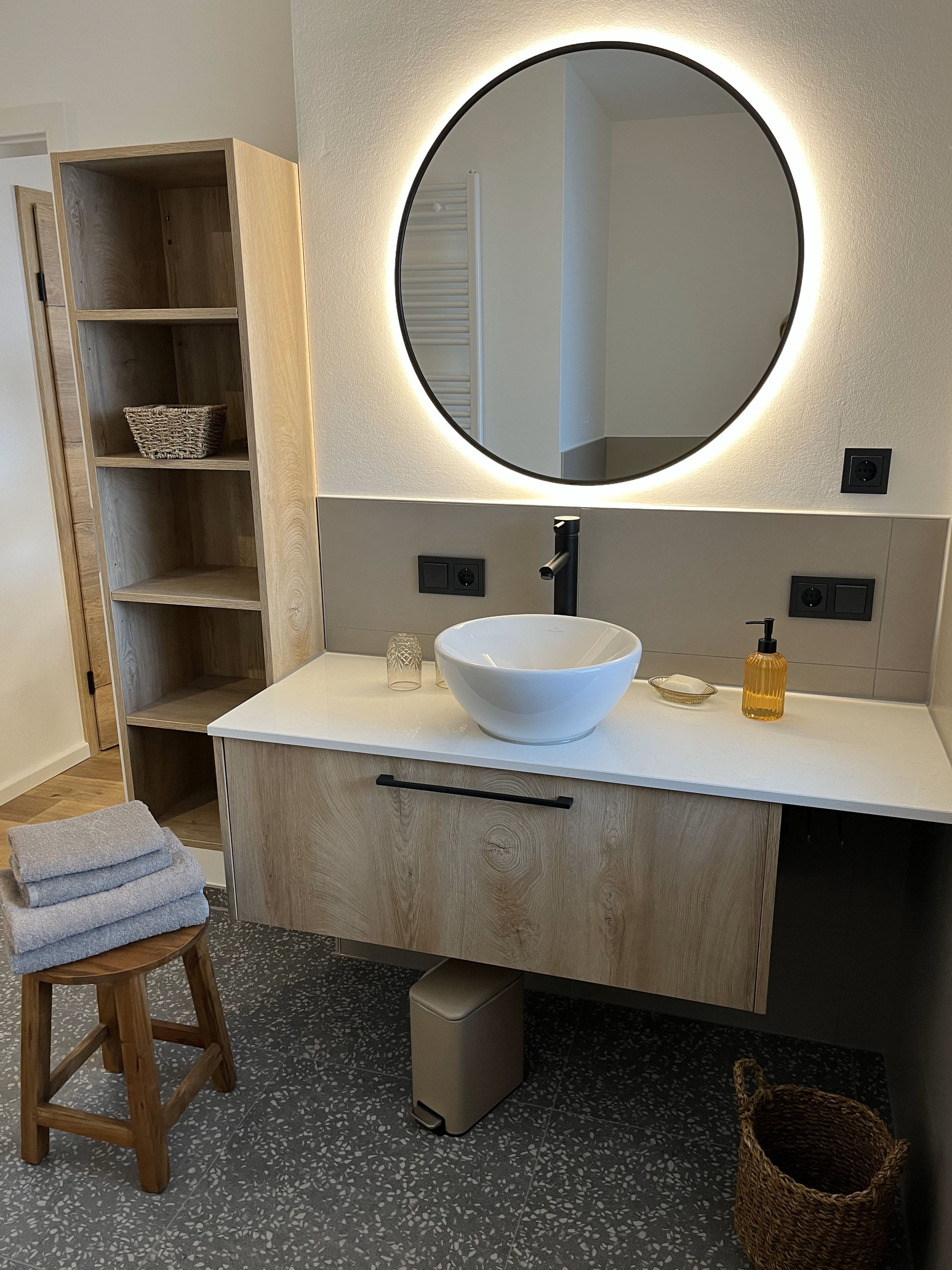 A bathroom with a white sink, a round mirror with a light, a wooden vanity, a stool with towels, and a tiled floor.