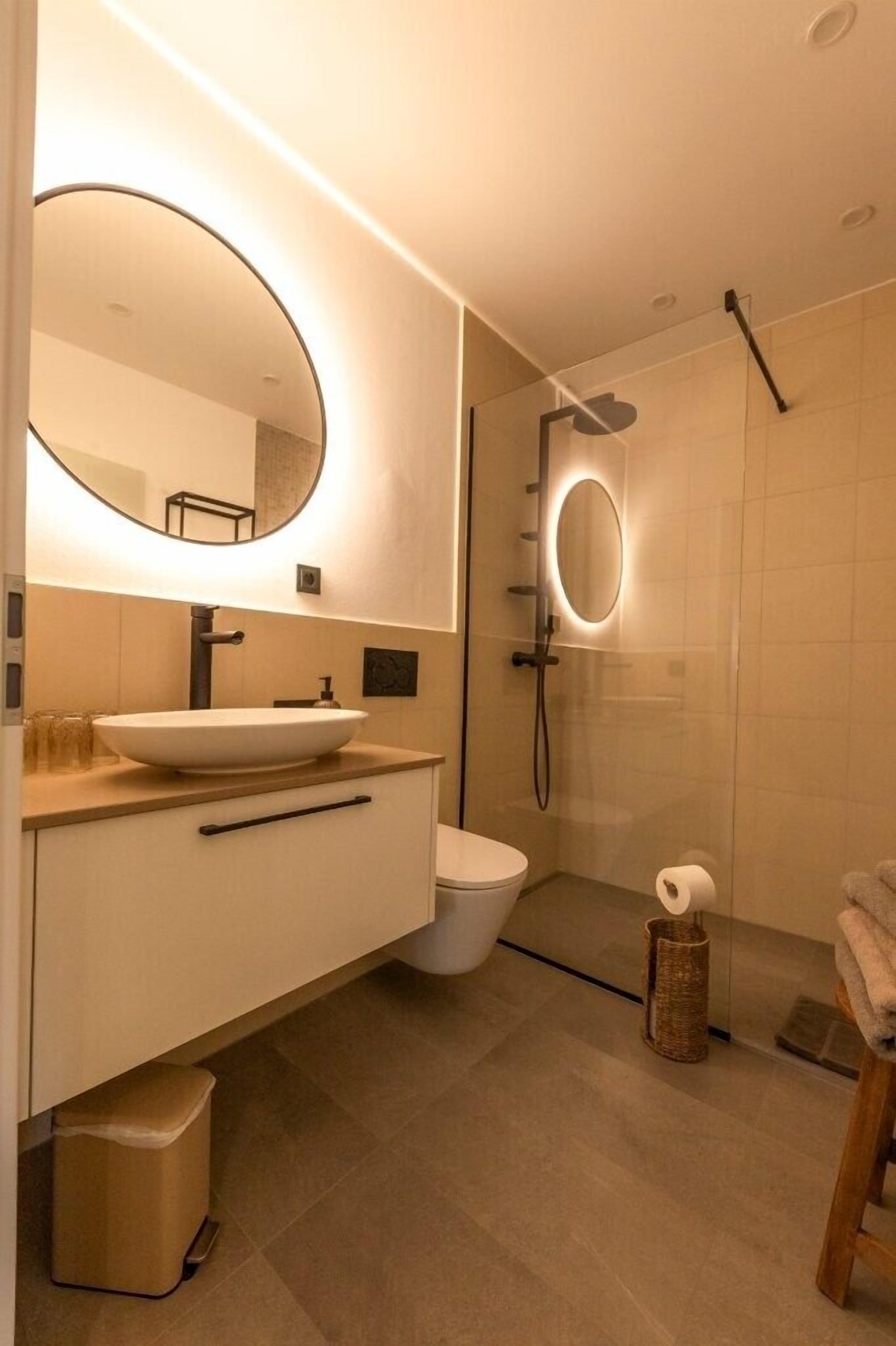 Une salle de bain moderne avec un miroir rond, un lavabo sur colonne, des toilettes et une douche à parois de verre.