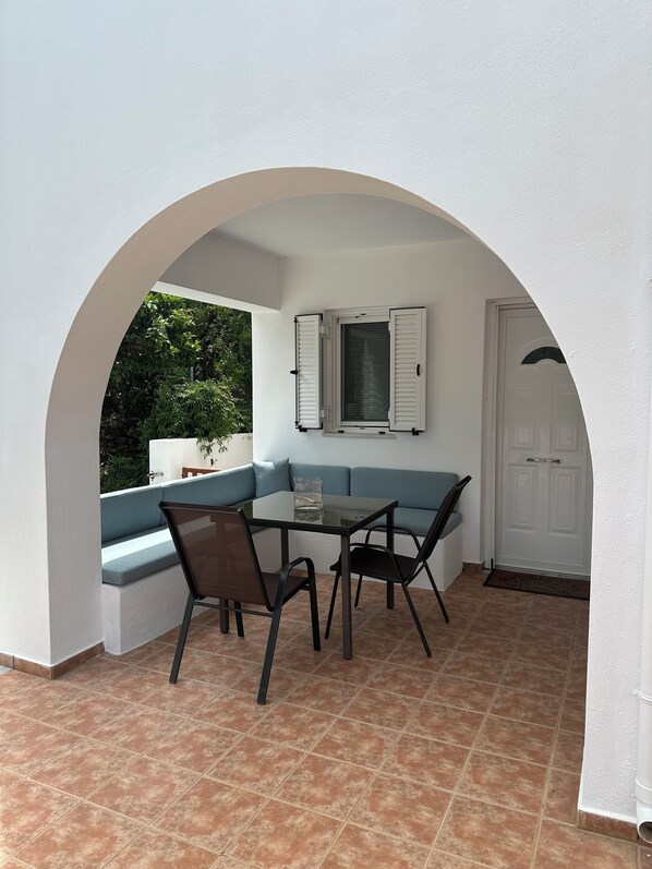 Een overdekt terras met een glazen tafel en stoelen, een blauwe, beklede bank, en een witte deur.