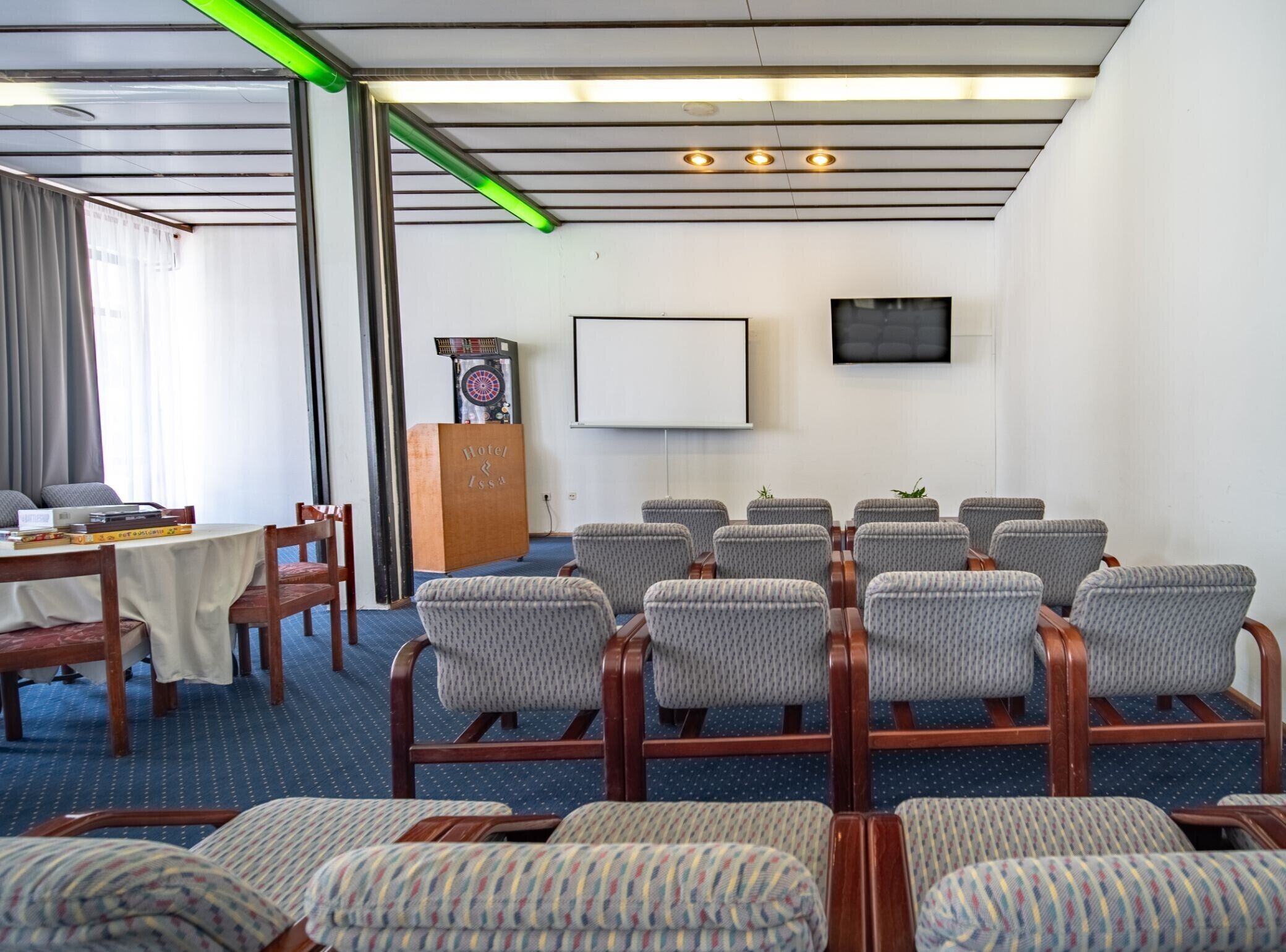 A conference room with a whiteboard, a podium, and a television screen. There are rows of chairs and a table with a laptop and books.