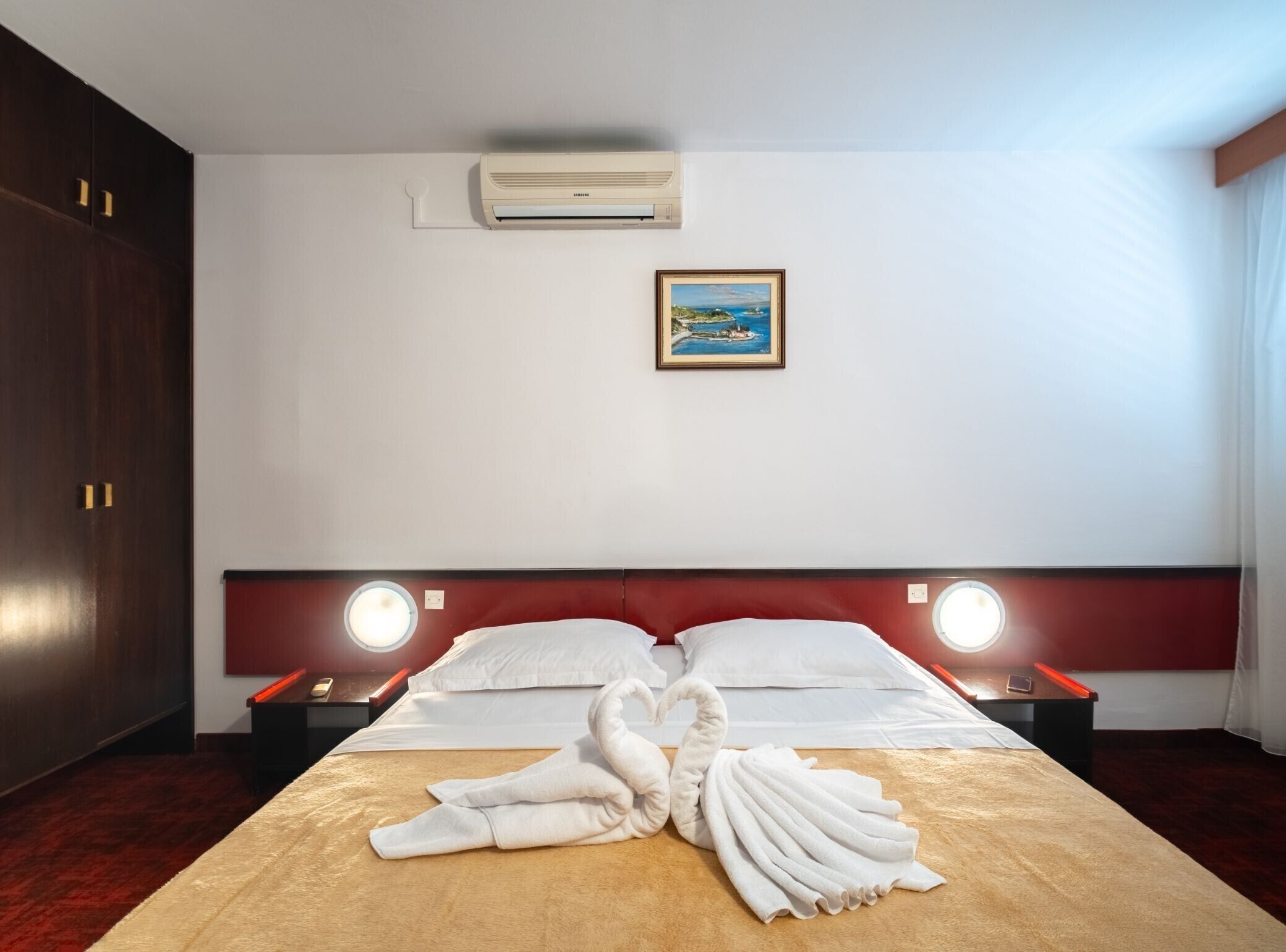 Une chambre d’hôtel comprenant un lit, deux tables de chevet, un climatiseur fixé au mur, un tableau et une décoration artistique réalisée avec des serviettes sur le lit.