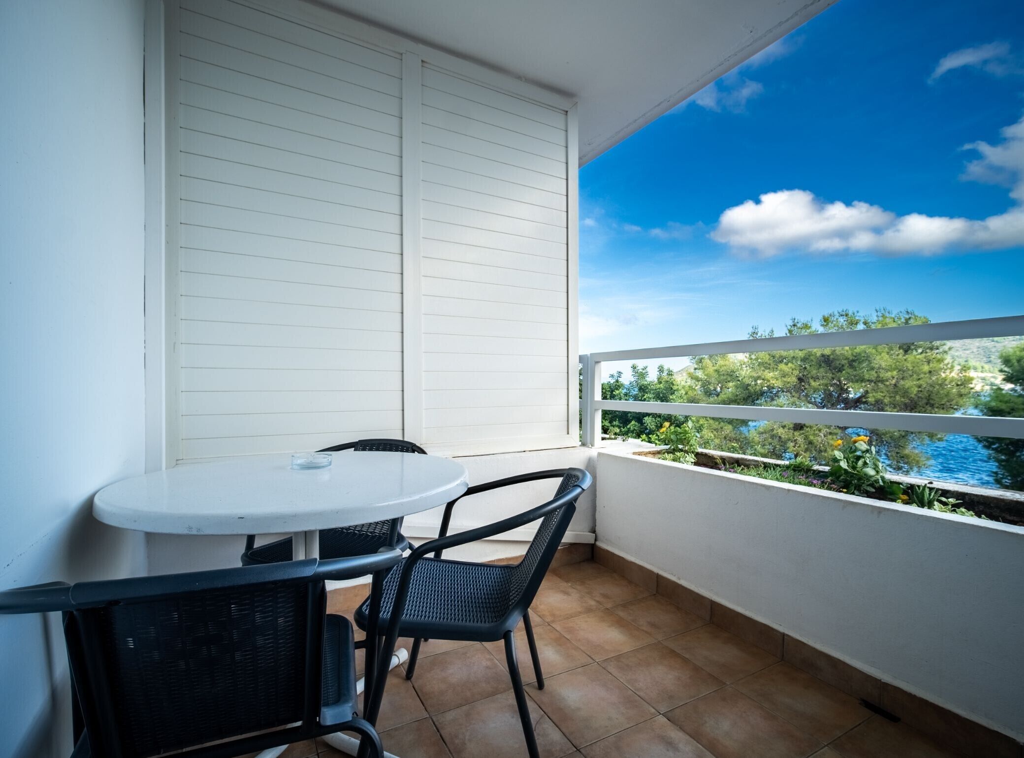 A balcony with a white table and two chairs, a view of greenery and water, and a clear blue sky.