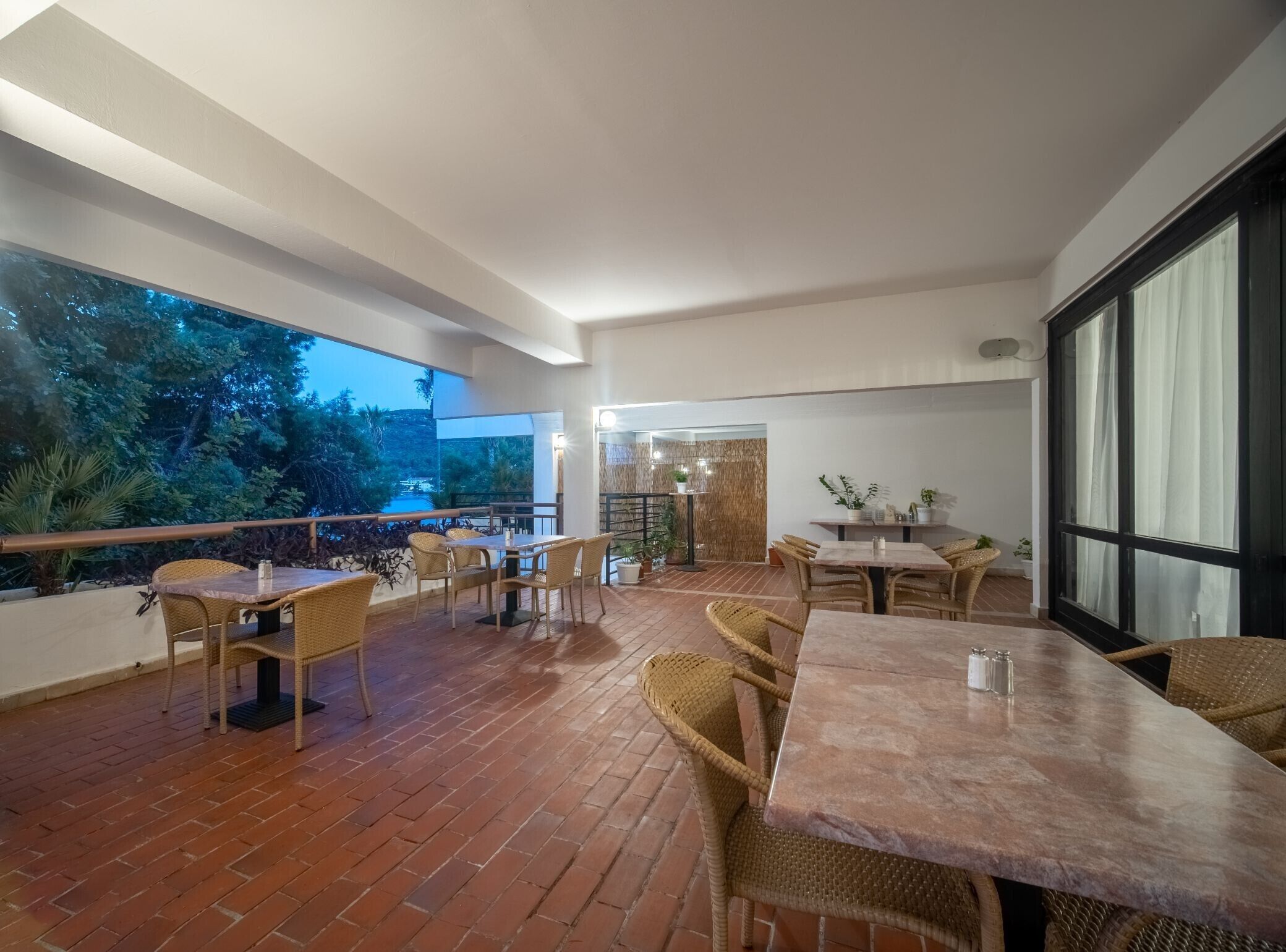 An outdoor patio area with brick flooring, wicker furniture, and large windows offering a view of trees and a distant landscape.
