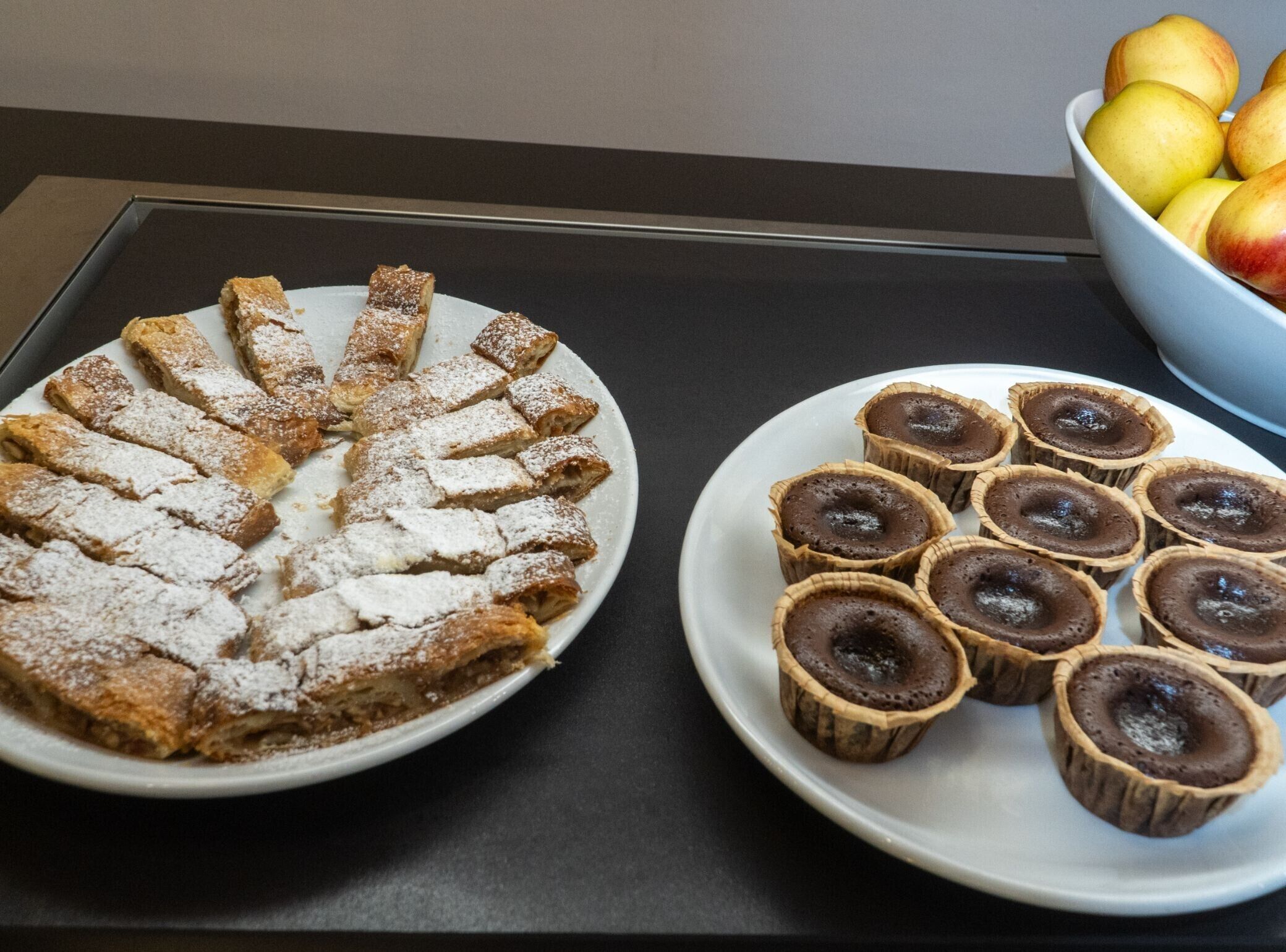 Un plato redondo y blanco con pasteles en rodajas espolvoreados con azúcar glas y una bandeja de tartaletas de chocolate.