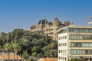 Un edificio histórico de varios pisos y una torre destacada, rodeado de abundante vegetación y edificios modernos.