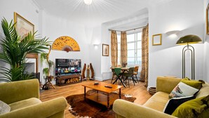 Ein Wohnzimmer mit Couch, zwei grünen Sesseln, einem Holztisch, einem Fernseher und einem Essbereich mit Tisch und Stühlen.