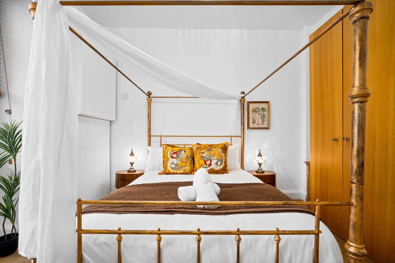A four-poster bed with a canopy, two bedside tables with lamps, a wooden wardrobe, and a potted plant.