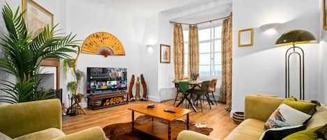 A living room with a wooden coffee table, two green armchairs, a sofa, a TV, and a dining area with a table and chairs.