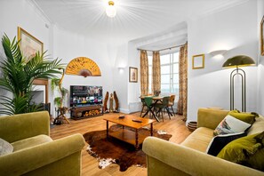 Ein Wohnzimmer mit Couch, zwei grünen Sesseln, einem Holztisch, einem Fernseher und einem Essbereich mit Tisch und Stühlen.