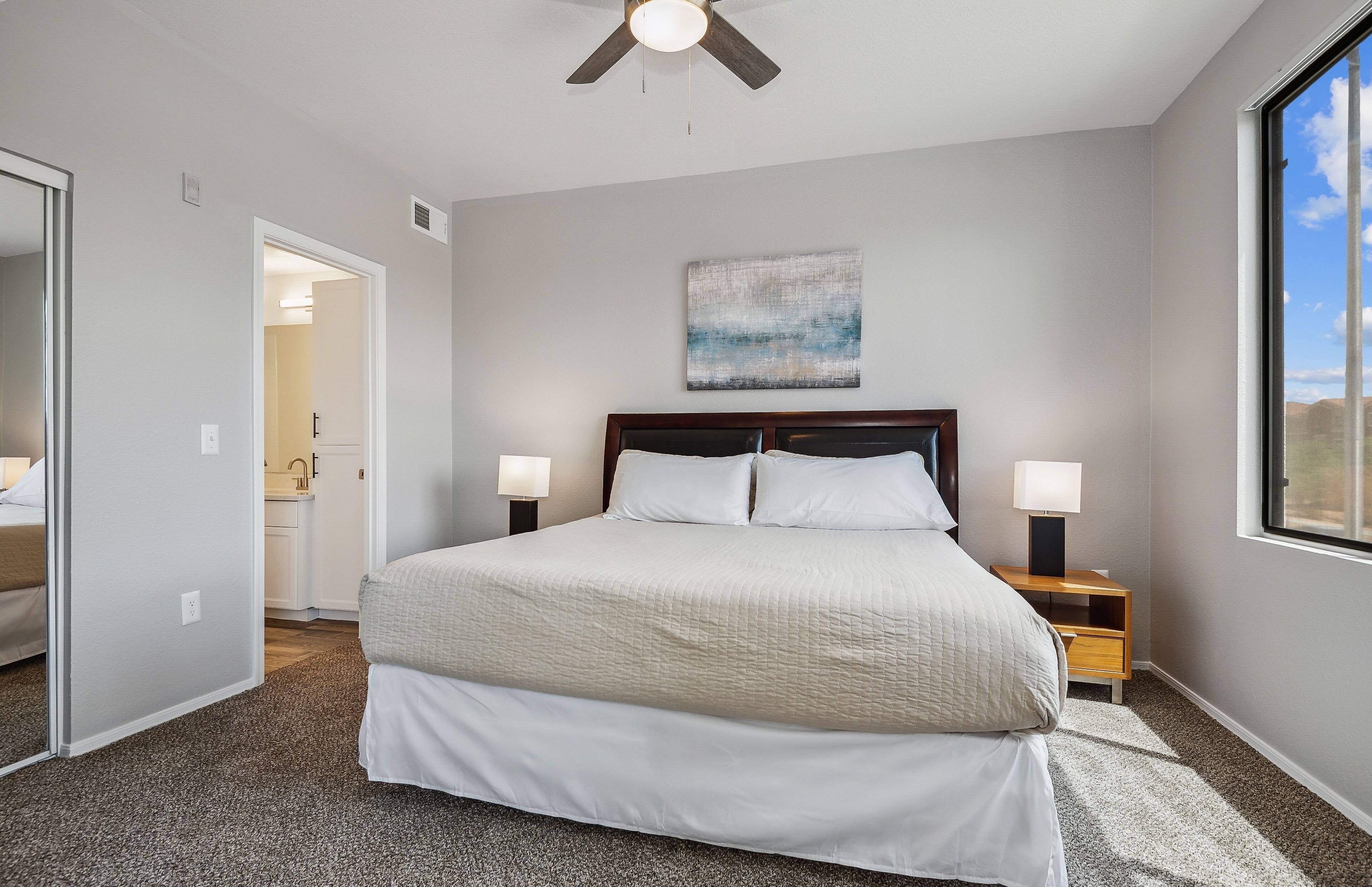 A bedroom with a large bed, two bedside tables with lamps, a ceiling fan, a window with a view, and a door leading to a bathroom.