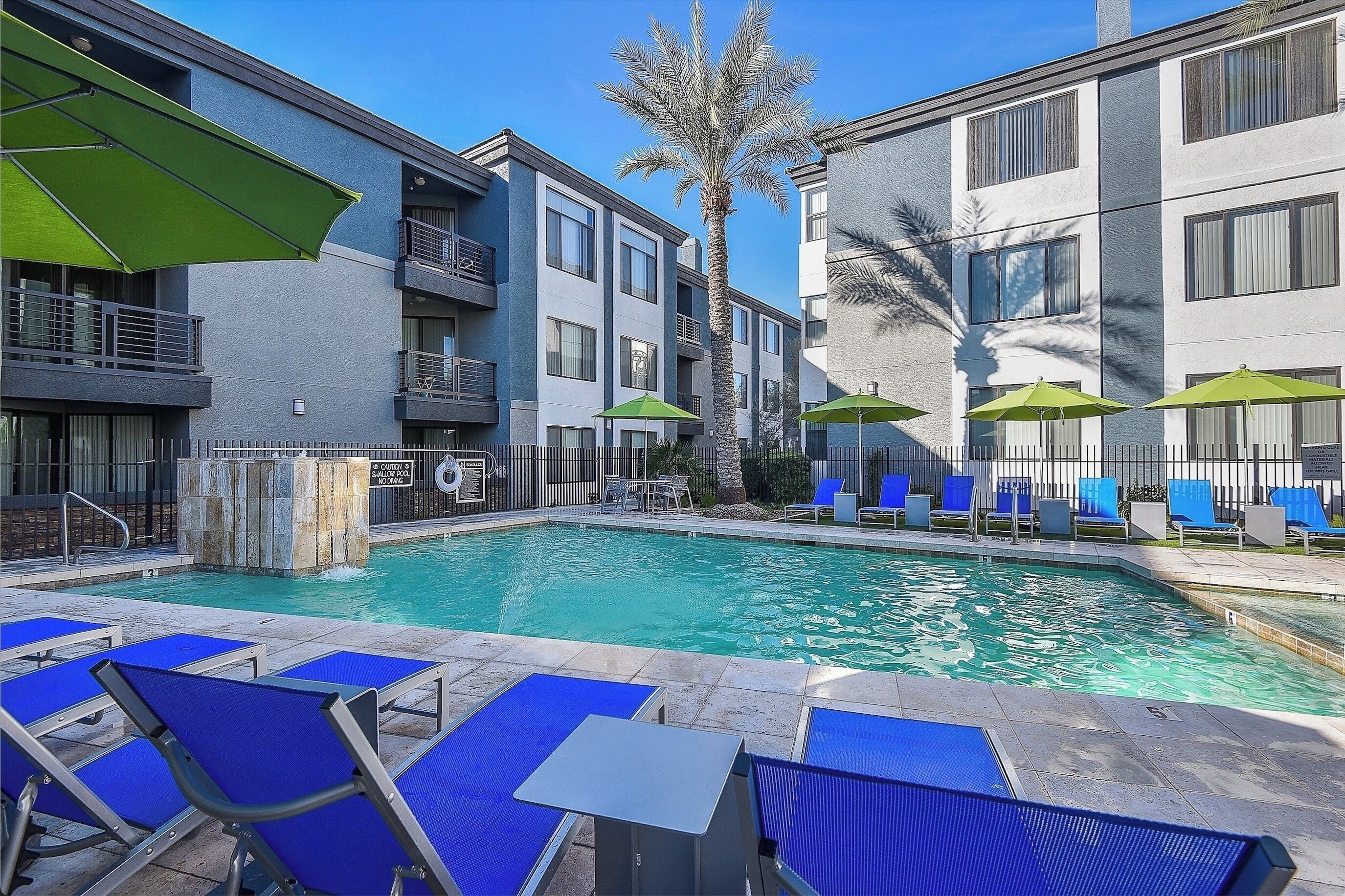 A swimming pool with blue lounge chairs and green umbrellas, surrounded by modern buildings.