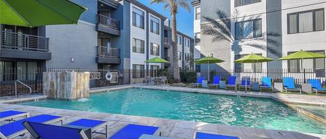 A swimming pool with blue lounge chairs and green umbrellas, surrounded by modern buildings.