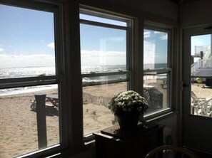 Habitación con grandes ventanales que dan a una playa, una planta en maceta sobre una mesa y vista al océano y muebles de playa.