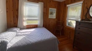 Una habitación con paneles de madera, una cama, un aparador y dos ventanas con cortinas.